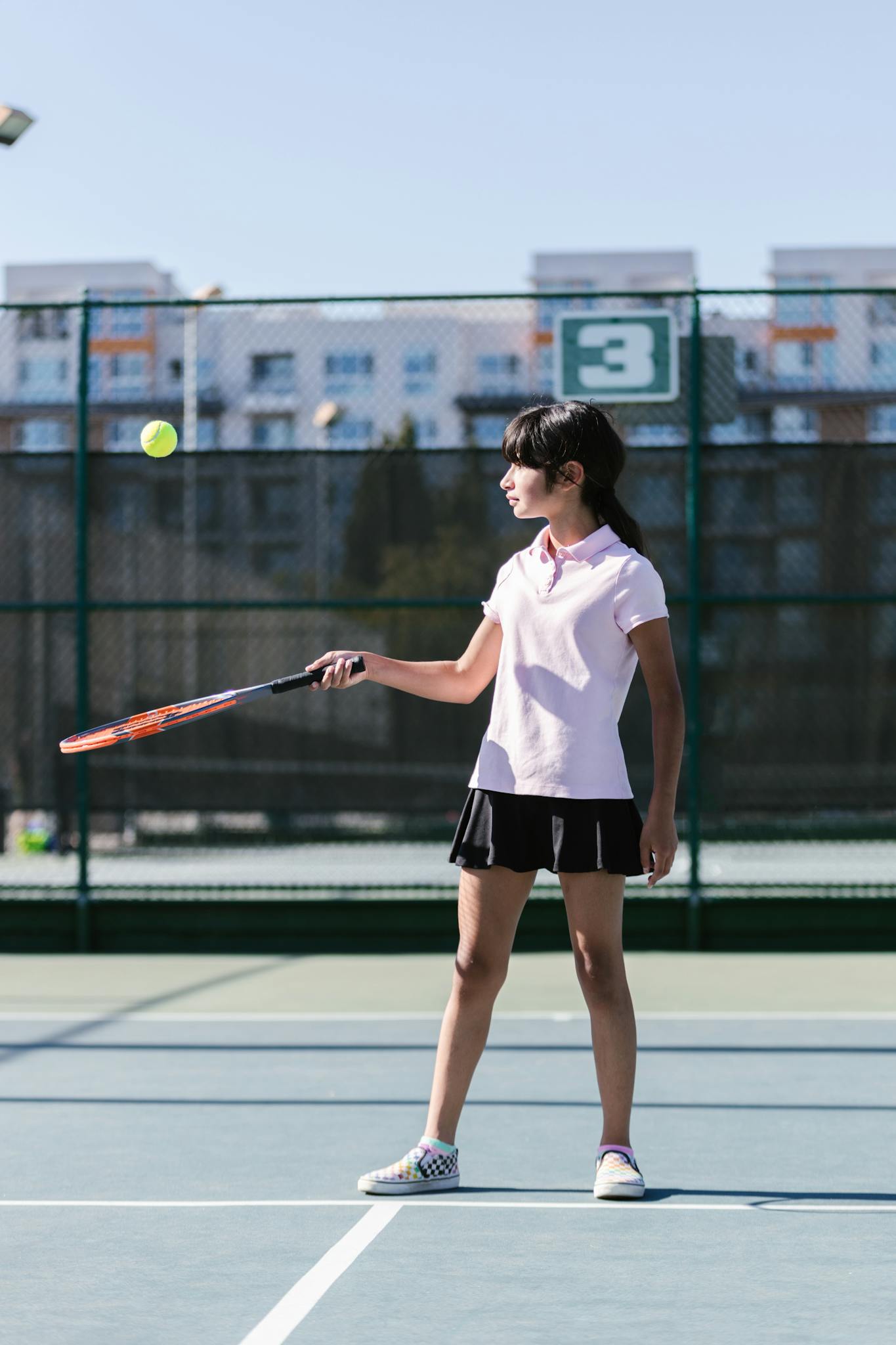 Teen girl practicing tennis with racket and ball on a sunny outdoor court.